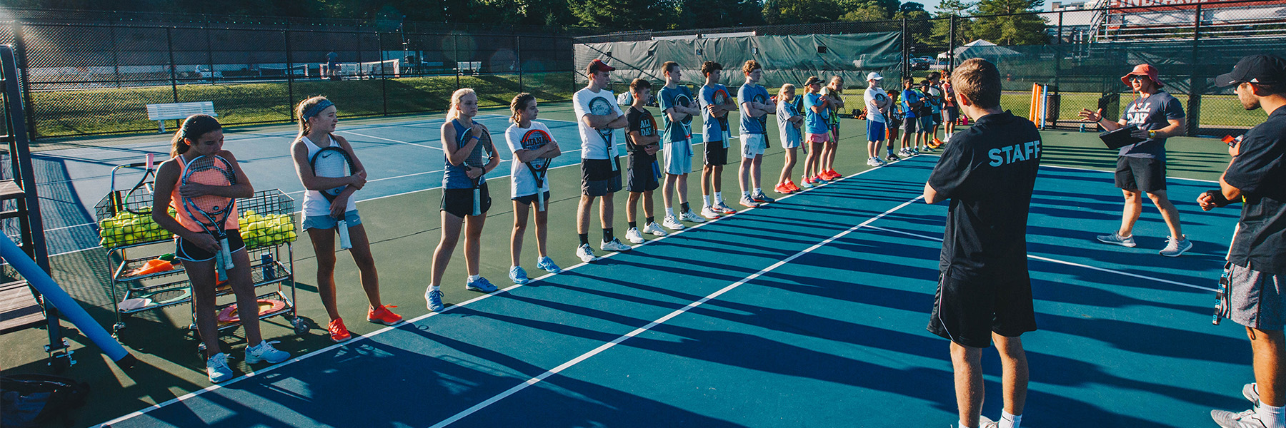 Three tennis instructors address a large group of junior players on an outdoor tennis court.